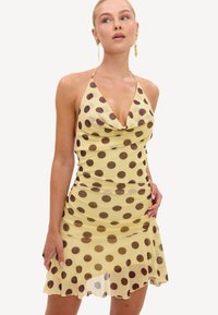 Femme portant une robe dos nu jaune avec de grands pois marron et un ourlet à volants, accessoirisée avec de longues boucles d'oreilles pendantes.