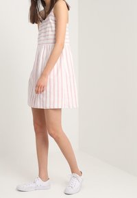 Vestido de verano de algodón a rayas con un corset ajustado, falda acampanada y tirantes finos en rosa y blanco. Combinado con zapatillas blancas.
