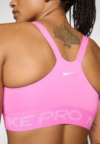 Rozā sporta krūšturis ar gludu tekstūru, platām siksnām un kontrastējošu balto logotipu. Iezīmēts ar ribotu joslu, kurā ir uzraksts "NIKE PRO" gaiši rozā krāsā.