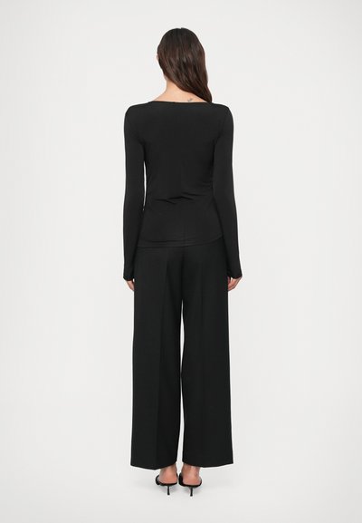 Hosszú sötét hajú nő, aki testhez simuló fekete, hosszú ujjú felsőt és bő szárú fekete nadrágot visel, mezítláb áll egy fehér háttér előtt.