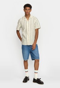 Korte mouwen, knoopsluiting shirt met crèmekleurige en blauwe strepen, gecombineerd met denim korte broek. Model draagt gestreepte sokken en zwarte schoenen.