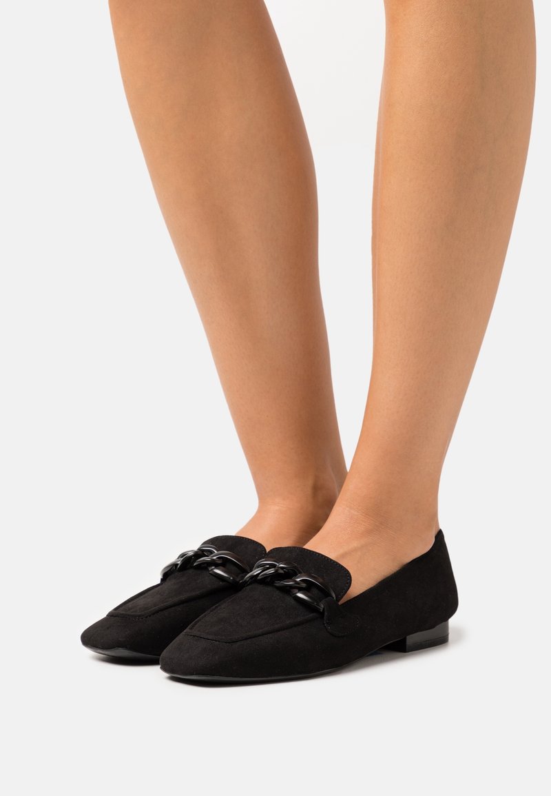 Jambes inférieures portant des mocassins en daim noir avec des bouts carrés arrondis et un détail décoratif en chaîne noire sur le dessus.