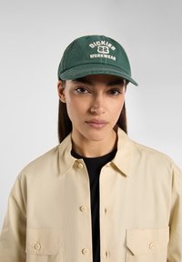 Vihreä puuvillalippis, jossa on kirjailtu valkoinen teksti "DICKIES 22 WORKWEAR." Omaaa kaarevan lippareunan ja ommellut yksityiskohdat. Henkilö, jolla on päällään beige paita, käyttää sitä.