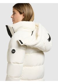 Balta pūkinė striukė su aukštu apykakliu, juodas logotipo lopas ir užtrauktuko detalės. Audinys lygus, su siuvinėta tekstūra.