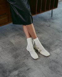 Chaussures en cuir verni blanc avec deux sangles et des boucles décoratives, portées avec des chaussettes blanches à côtes, reposant sur un sol gris texturé.