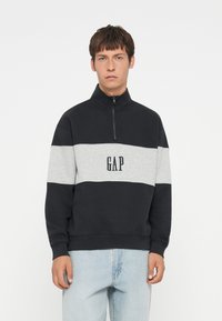 Čierna a šedá farebne rozdelená mikina s vysokým golierom a zipsom. Obsahuje čierne logo "GAP", vyrobené z mäkkej textúrovanej látky.