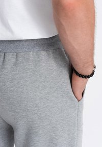 Grijze katoenen joggingbroeken met een ribbelband, zijzakken en een subtiele textuur. Een zwart kralenarmband is zichtbaar om de pols.