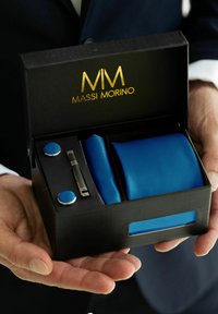 Ensemble cravate bleue présenté dans une boîte noire avec inscription dorée, comprend des boutons de manchette assortis et une pince à cravate. Texture lisse, design minimaliste et présentation élégante.