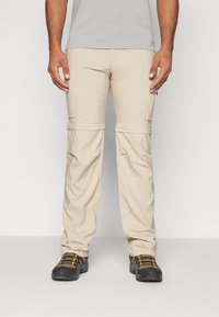 Pantalones cargo beige con cremalleras desmontables y bolsillos con cremallera, de corte relajado, combinados con zapatos de senderismo negros con detalles en naranja.