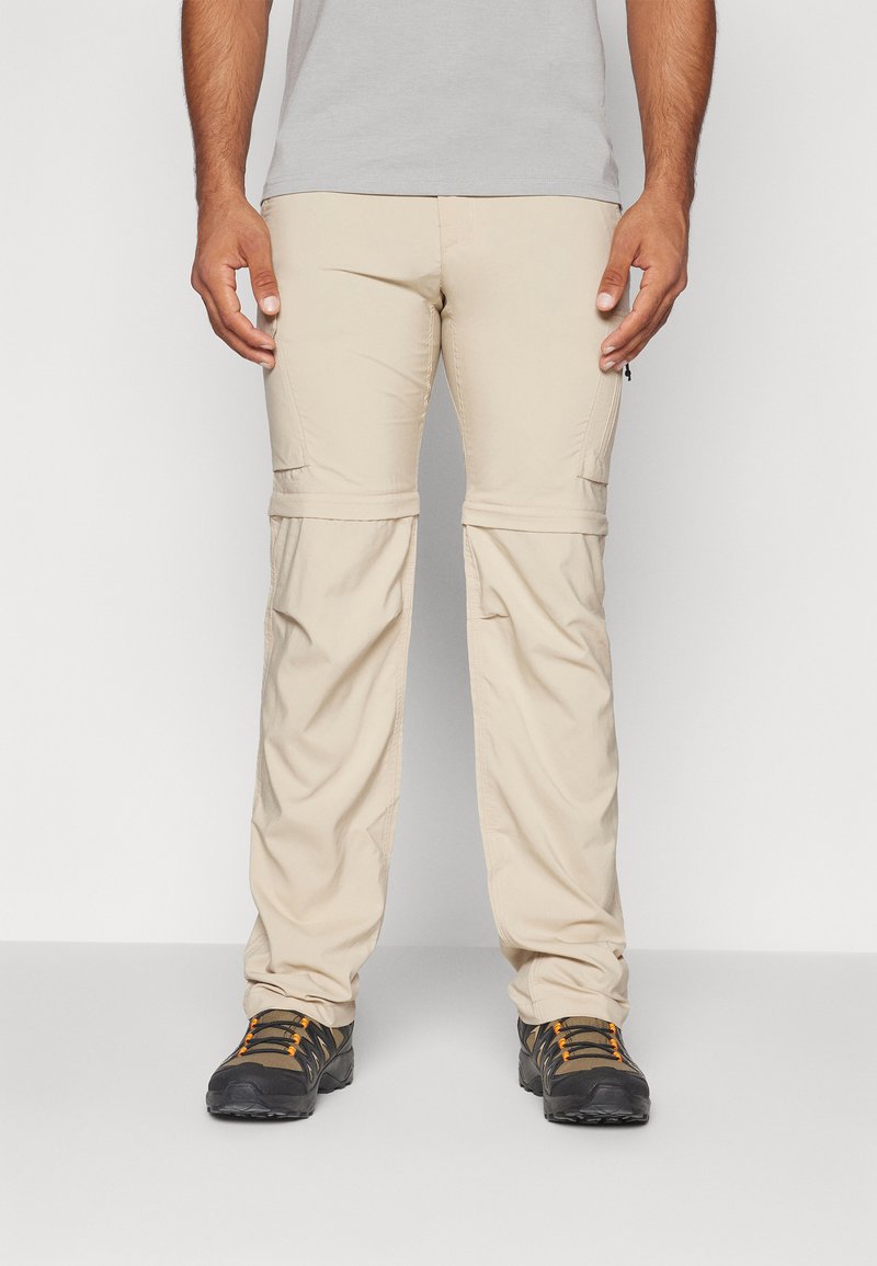 Pantalones cargo beige con cremalleras desmontables y bolsillos con cremallera, de corte relajado, combinados con zapatos de senderismo negros con detalles en naranja.