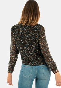 Long-sleeve, sheer black blouse with small floral pattern, featuring a gathered neckline and elastic cuffs, paired with light blue jeans.