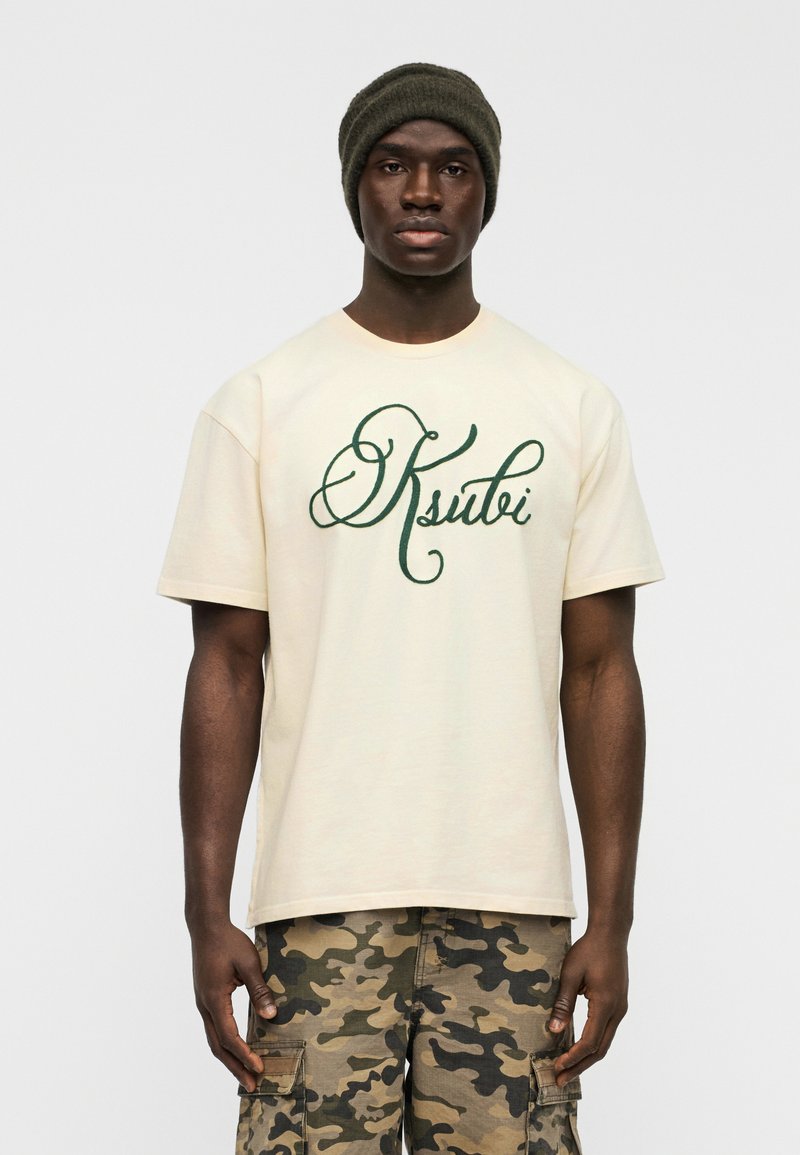 Κοντομάνικη κρεμ μπλούζα με πράσινο καλλιγραφικό λογότυπο "Ksubi". Συνδυασμένη με παραλλαγής cargo σορτς. Το μοντέλο φοράει σκουρό πράσινο σκουφί.