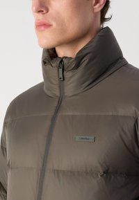 Veste matelassée gris foncé avec un col haut, fermeture éclair à l'avant et une texture légèrement brillante. Présente une petite étiquette logo sur la zone du poitrail gauche.