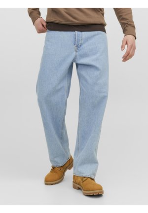 Personne portant un jean bleu clair ample, des bottes marron et un haut marron à manches longues, debout avec une main près de la poche du jean.