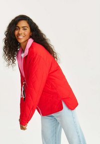 Červená prešívaná bunda s ružovými detailmi, obsahujúca zips, vrecká a bočné rozparky. Model nosí svetlo modré džínsy.