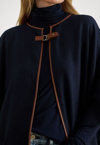 Tamsiai mėlynas megztas viršutinis drabužis su zigzaguota tekstūra, rudo odos apdaila ir užsegimu. Dėvimas ant tamsaus golfų.