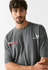 Szürke, rövid ujjú póló puha anyagból, fehér "Jeep" és piros "WRANGLER" logókkal, kényelmes szabással és kerek nyakkivágással.