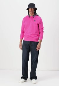 Sudadera fucsia con cuello redondo, mangas largas y un pequeño logotipo de la marca, combinada con pantalones de mezclilla oscuros y zapatillas blancas.