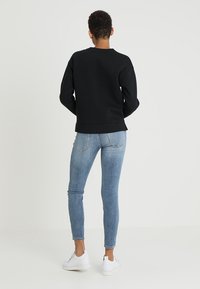 Czarna bluza z okrągłym dekoltem i ściągaczami na rękawach, zestawiona z jasnoniebieskimi obcisłymi jeansami. Model nosi białe trampki, prezentując casualowy wygląd.