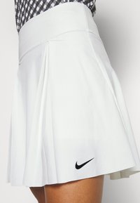 Bílá skládaná sukně vyrobená z lehkého materiálu, s širokým pasem a malým černým logem Nike v pravém dolním rohu.