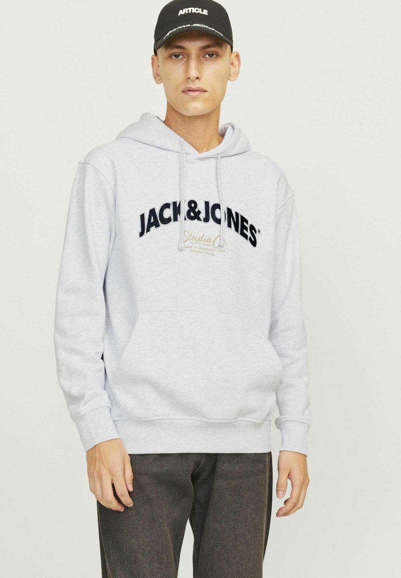 Grauer Kapuzenpullover mit einer Fronttasche und Kordeln, ausgestattet mit einem schwarzen Logo "JACK & JONES" und "Studio C" in Gold. Getragen mit dunklen Hosen.