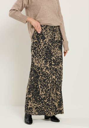 Person trägt einen beigen Pullover und einen langen Rock mit Leopardenmuster sowie schwarze Stiefeletten, steht mit einer Hand in der Rocktasche.