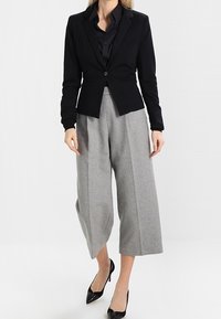 Kvinna som har på sig en svart figurnära kavaj, svart skjorta, vida grå cropped-byxor och svarta spetsiga klackar, står mot en vit bakgrund.