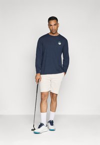 Košulja dugih rukava tamnoplave boje s logom sprijeda, bež šortsevi, bijele čarape i golf cipele s plavim detaljima. Drži golf štap. Čist, suvremen izgled.