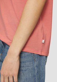 Różowa koszulka z krótkim rękawem, uszyta z miękkiego materiału, z okrągłym dekoltem i małą metką marki z boku, zestawiona z dżinsami.