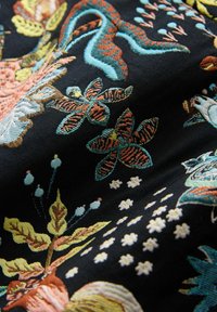 Tissu noir avec des motifs floraux et aquatiques vibrants dans des tons de bleu, rose, jaune et orange, présentant une broderie texturée et des détails complexes.