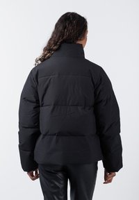 Schwarze Daunenjacke mit hohem Kragen, gestepptem Design und lockerer Passform. Das Material wirkt glatt, ohne sichtbare Muster oder Akzente.