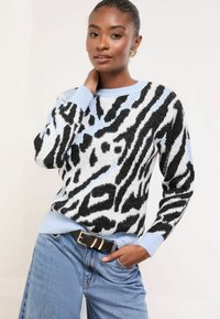 Kötött pulóver kék és fekete zebra mintával, puha textúrával, kerek nyakkal és bordázott szegéllyel. Magas derekú világos farmernadrággal párosítva.
