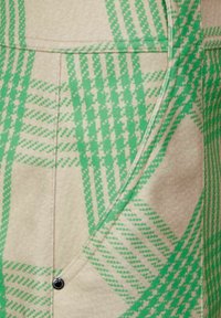 Beige Stoff mit grünem Karomuster, mit schrägen Nähten und einem schwarzen Druckknopf-Akzent. Strukturierte Oberfläche mit kontrastierenden Farben.