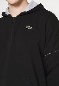 Homme portant un sweat à capuche noir avec demi-fermeture éclair, capuche intérieure grise et petit logo crocodile vert sur la poitrine.
