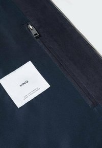 Tessuto blu navy con una texture liscia, dotato di una tasca con zip e un'etichetta bianca con la scritta "MNG" in rilievo. È visibile la ferramenta della cerniera argento.