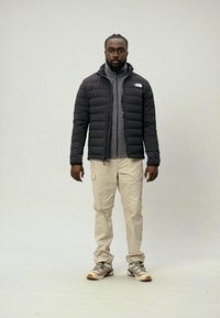 Casaco puffer preto com fecho na frente, camada interior cinza, calças cargo creme e sapatilhas claras sobre um fundo neutro.