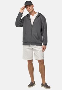 Sudadera gris de cremallera con bolsillos, llevada sobre una camiseta blanca de cuello en V, combinada con pantalones cortos claros y zapatos gris de tipo slip-on. La gorra combina con la sudadera.