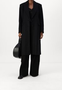 Sort dobbeltbrystet frakke med brede klapper, parret med løse sorte bukser og en struktureret sort håndtaske, alt præsenteret i en glat, elegant fremtoning.