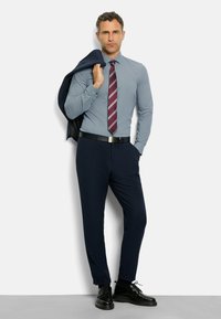 Mann in einem gemusterten blauen Hemd, gestreifter roter Krawatte, marineblauen Hosen, schwarzen Schuhen, der eine marineblaue Jacke über der Schulter trägt und vor einem weißen Hintergrund steht.