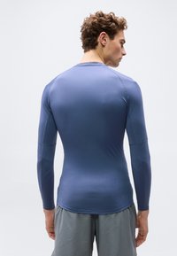 Νεαρός άνδρας με σγουρά μαλλιά που φοράει στενή μπλε αθλητική μπλούζα με μακριά μανίκια και γκρι σορτς, ο οποίος φαίνεται από την πίσω πλευρά με φόντο απλή επιφάνεια.