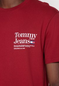 T-shirt en coton rouge avec un logo blanc affichant "Tommy Jeans" et des noms de villes. Manches courtes et col rond classique. Texture douce.