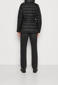 Czarna kurtka puchowa z poziomym pikowaniem, elastycznym pasem, wysokim kołnierzem i detalem logo na szyi, zestawiona z ciemnymi dżinsami i botkami.