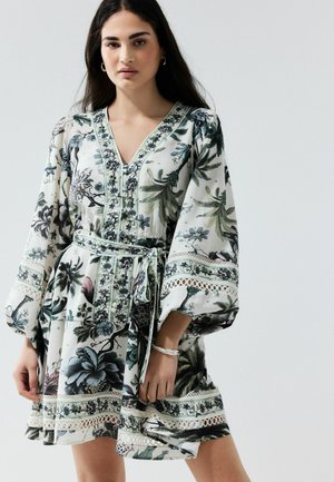Kvinde iført en bæltebeklædt, langærmet, blomsterprintet kjole med V-hals og blonderkant, stående mod en ensfarvet lys baggrund.