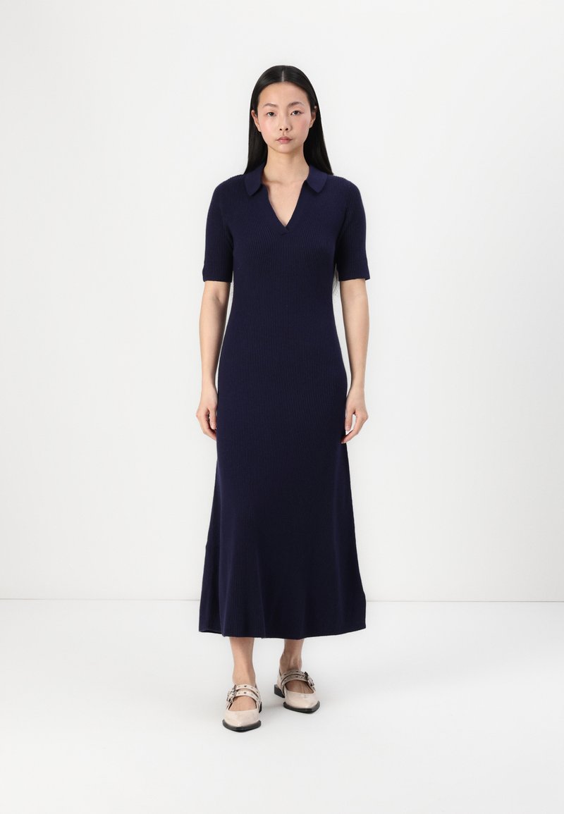 Navy ribbed midi dress with short sleeves and a V-neck collar, featuring a flared hem. Paired with neutral-toned flat shoes.