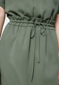 Robe vert olive avec une taille élastique froncée et un lien, confectionnée dans un tissu lisse et léger ; silhouette ample et décontractée.