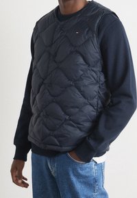 Navy Steppweste mit Rautenmuster und Druckknöpfen, über einem navy Sweatshirt getragen, kombiniert mit blauen Jeans.
