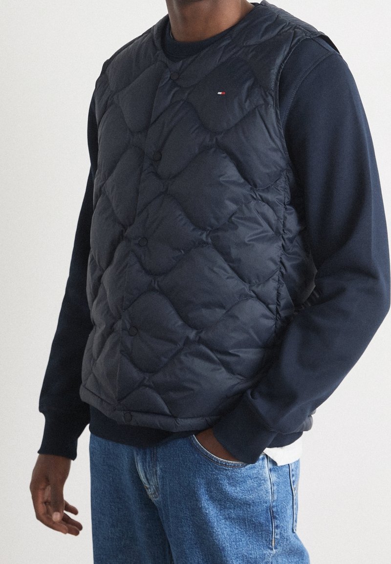 Navy Steppweste mit Rautenmuster und Druckknöpfen, über einem navy Sweatshirt getragen, kombiniert mit blauen Jeans.