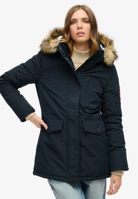 Navy puffer jacket with a fur-lined hood, zippered and button closures, and side pockets, worn over a light-colored sweater and jeans.