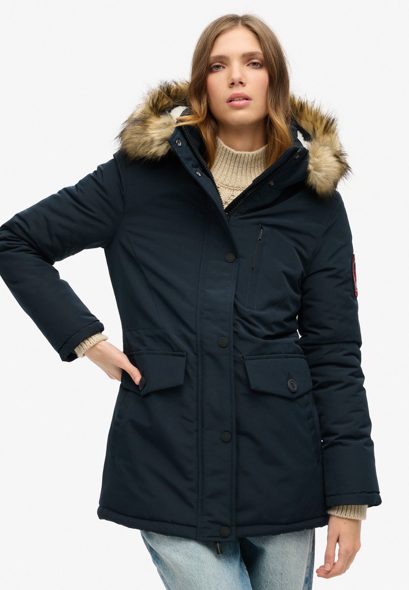 Navy puffer jacket with a fur-lined hood, zippered and button closures, and side pockets, worn over a light-colored sweater and jeans.