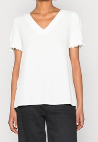 Biała bluzka z krótkimi rękawami, z dekoltem w kształcie V i zgromadzonymi falbanami na rękawach; wykonana z lekkiego materiału o gładkiej fakturze.